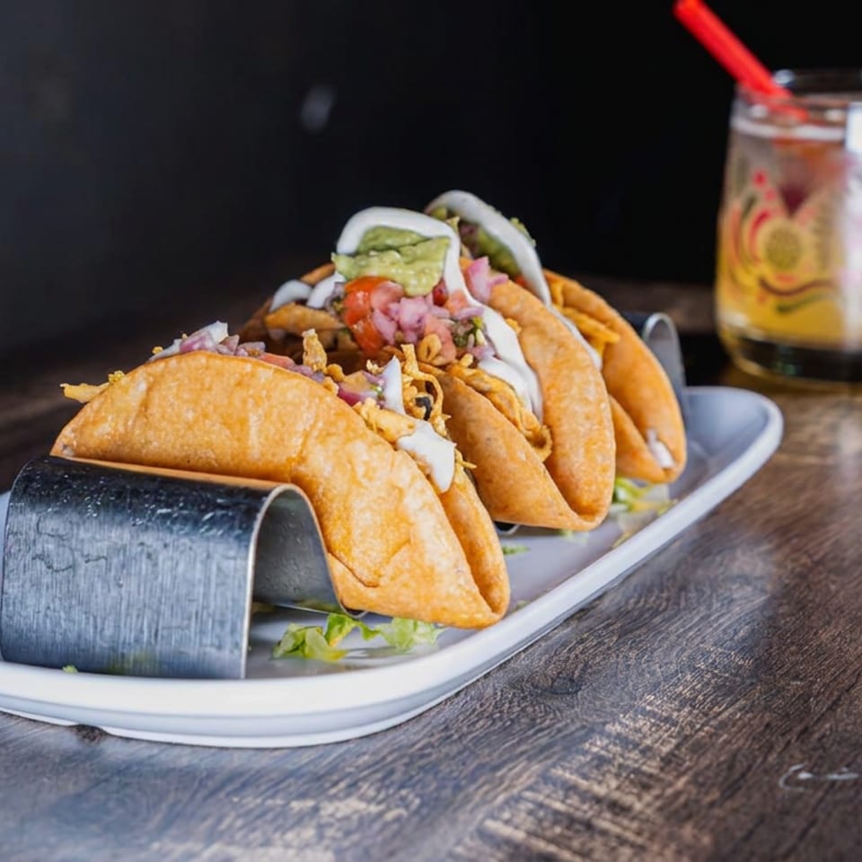 Crispy tacos topped with guacamole, pickled onions, and crema on a platter beside a colorful drink with a red straw.