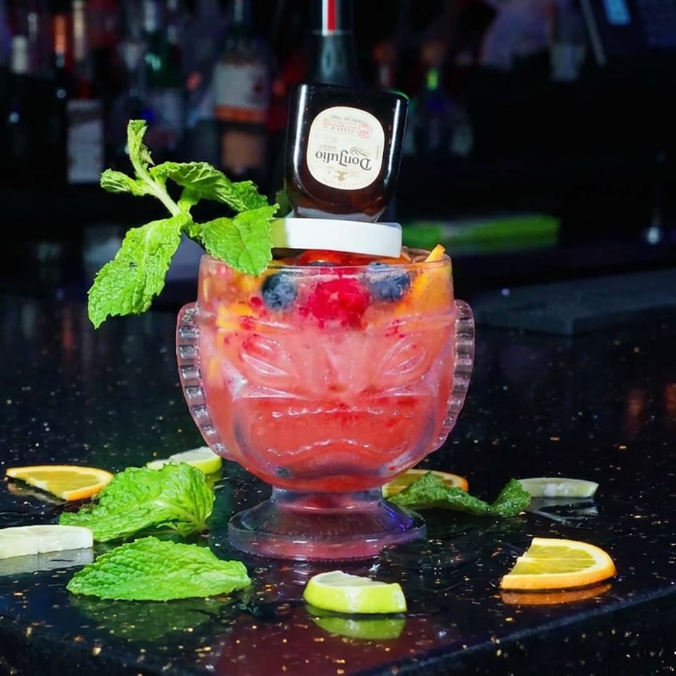 Red fruit cocktail with mint, citrus garnishes, and an inverted liquor bottle served on a reflective bar counter.