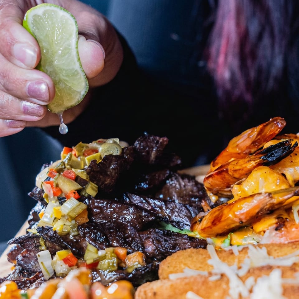 Person squeezes lime over a platter of grilled shrimp, dark meat, vegetables, and golden fried sides.