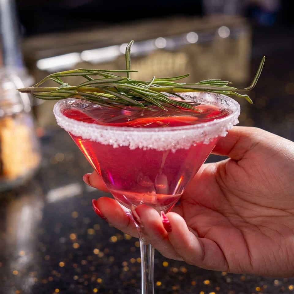 Hand holding a bright red cocktail in a sugar-rimmed martini glass garnished with fresh rosemary at a dimly lit bar.