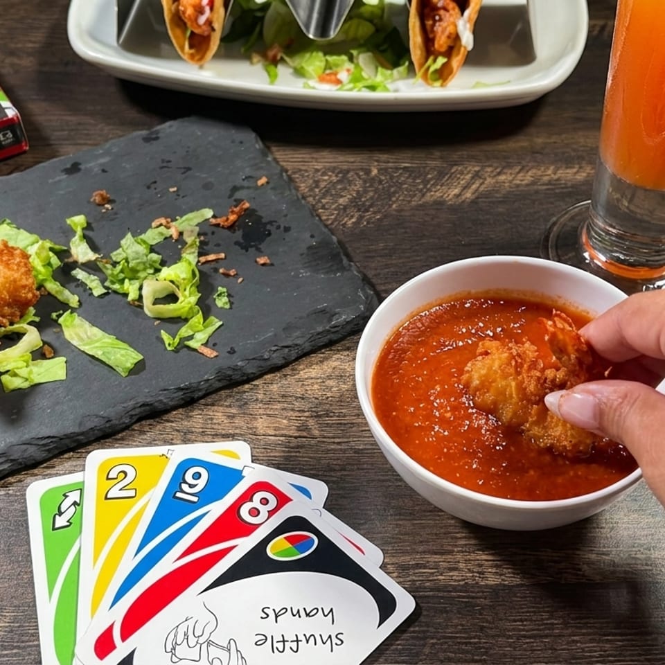 A hand dips a crispy fried appetizer into red sauce beside lettuce, tacos, an orange drink, and UNO cards on a wooden table.