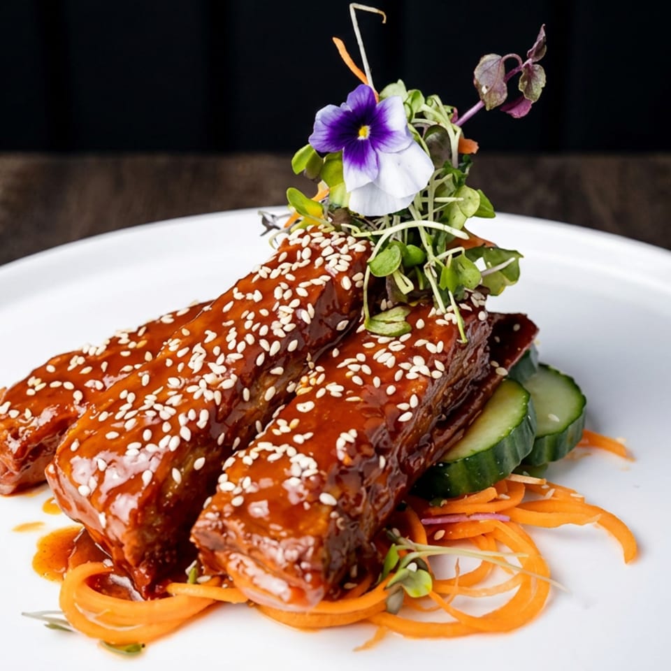 Glazed sesame-crusted ribs served with cucumber, shredded carrots, microgreens, and edible flowers on a white plate.