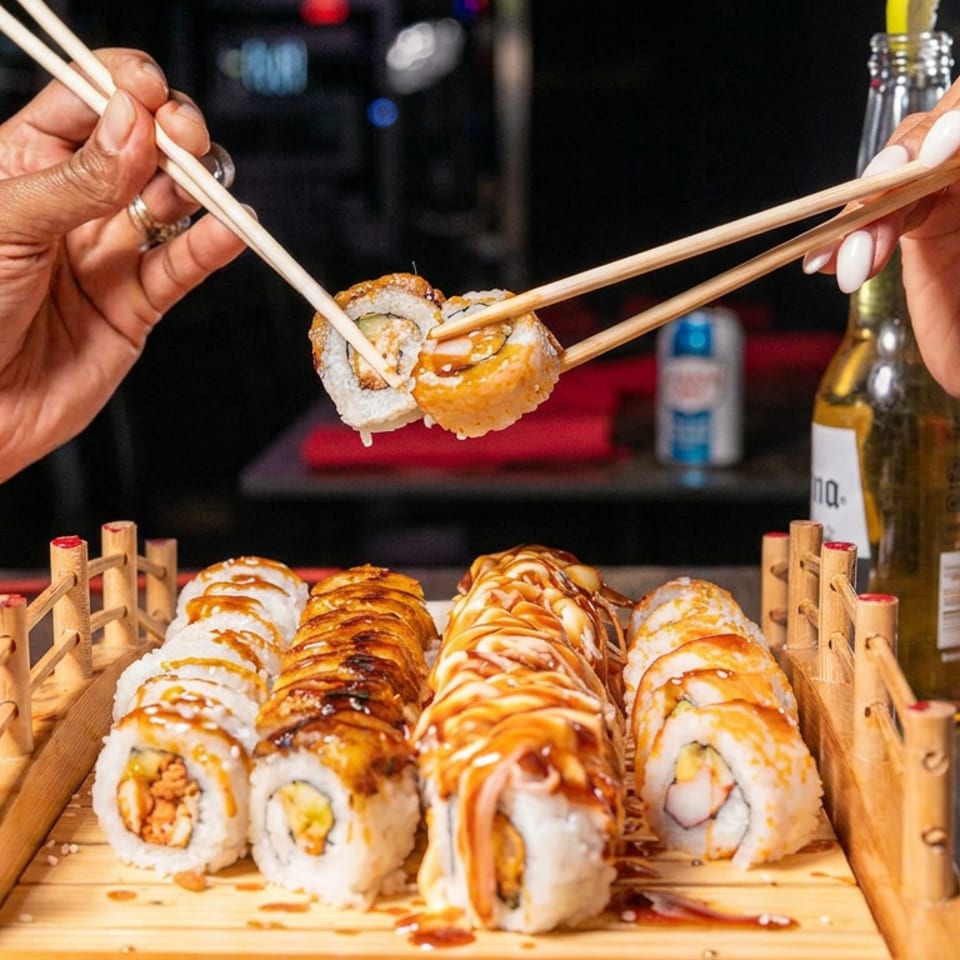 Hands use chopsticks to serve sushi rolls from a wooden platter in a dimly lit restaurant.