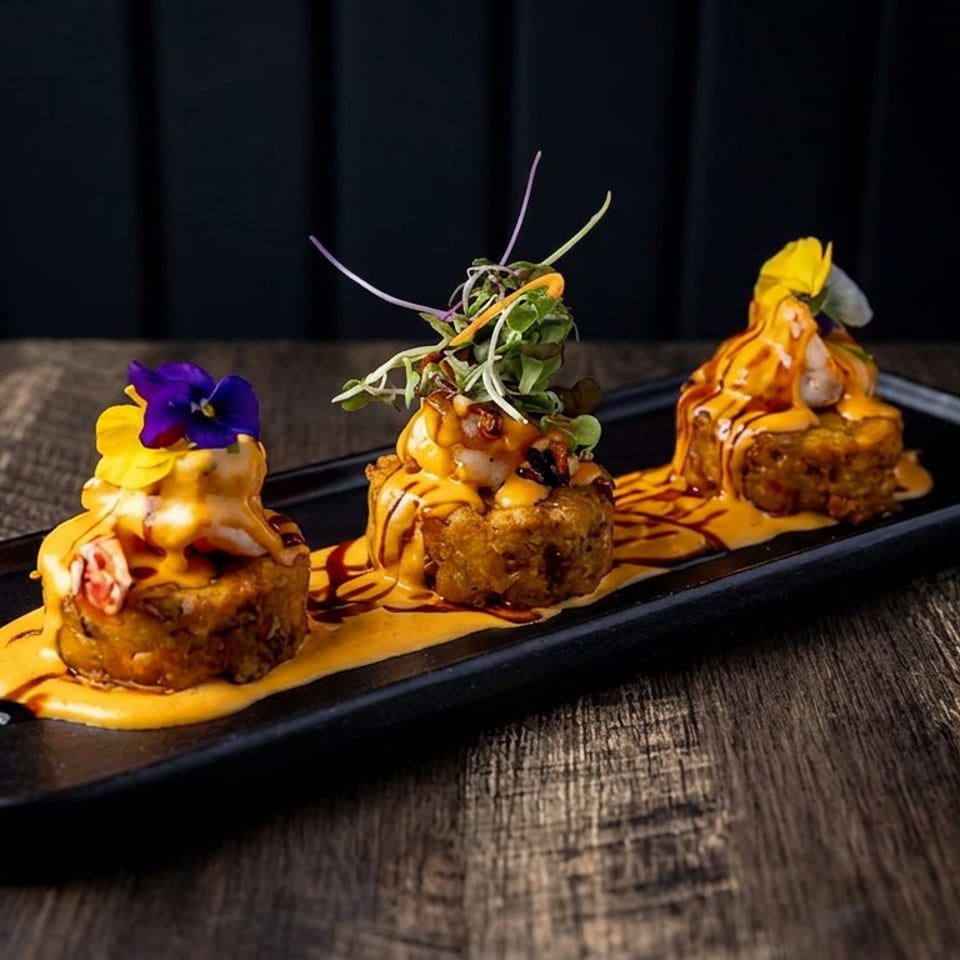 Three golden-brown savory bites topped with orange sauce, microgreens, and edible flowers on a black rectangular plate.