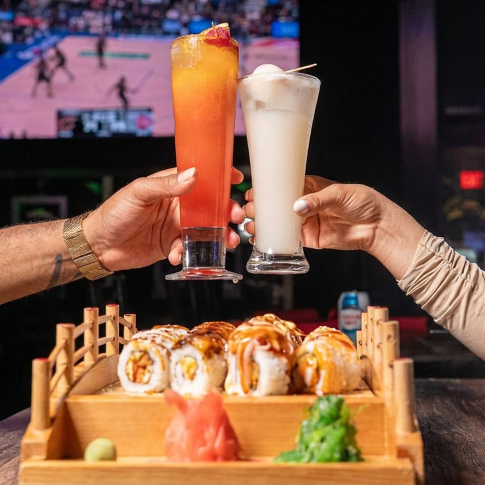 Hands toast colorful cocktails above a wooden tray of sushi rolls in a sports bar with a basketball game on television.