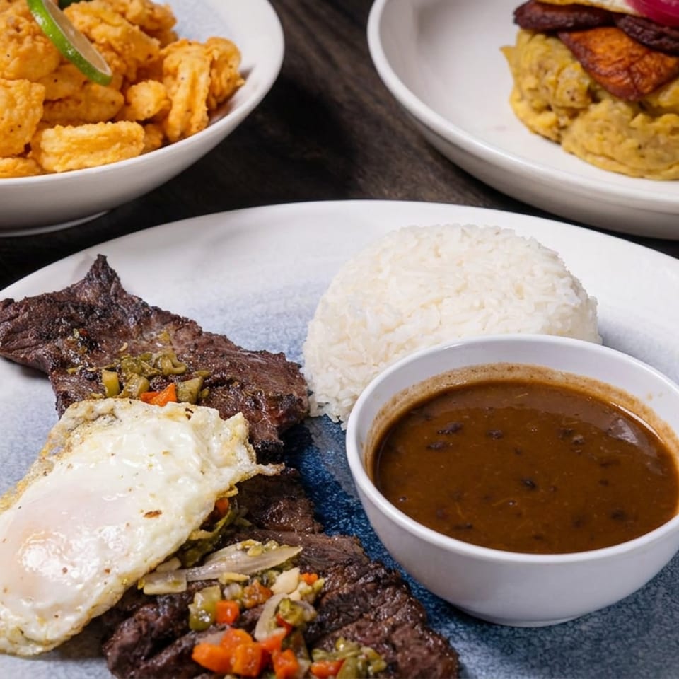 Plate of grilled steak topped with a fried egg, white rice, sautéed vegetables, and brown gravy, with fried seafood and a sandwich nearby.